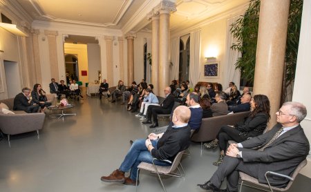 Persone sedute in una grande stanza con colonne che ascoltano una discussione su divani