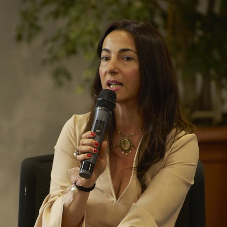 Donna con capelli lunghi e scuri parla al microfono, seduta con gambe incrociate su sedia