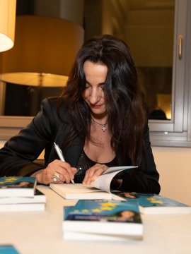Donna con capelli scuri firma libri su un tavolo con lampada e bicchiere di vino